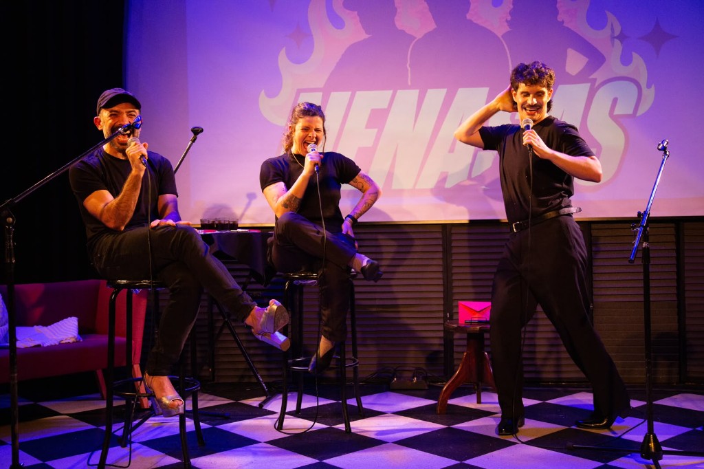 Los tres de Nenasas en vivo, con micrófono, frente al logo proyectado